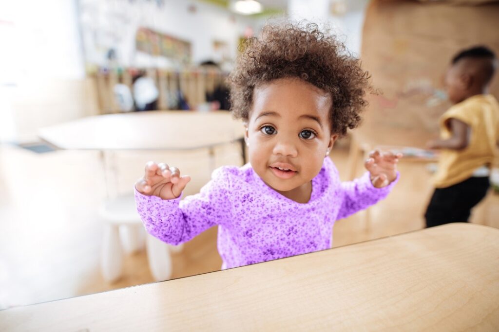 A toddler wearing a purple onesie standing beside a table with her hands up 
