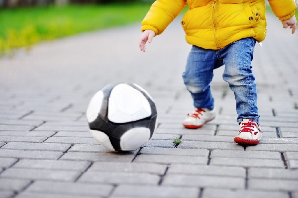  A toddler wearing a yellow jacket, jeans and rubber shoes kicking a soccer ball 