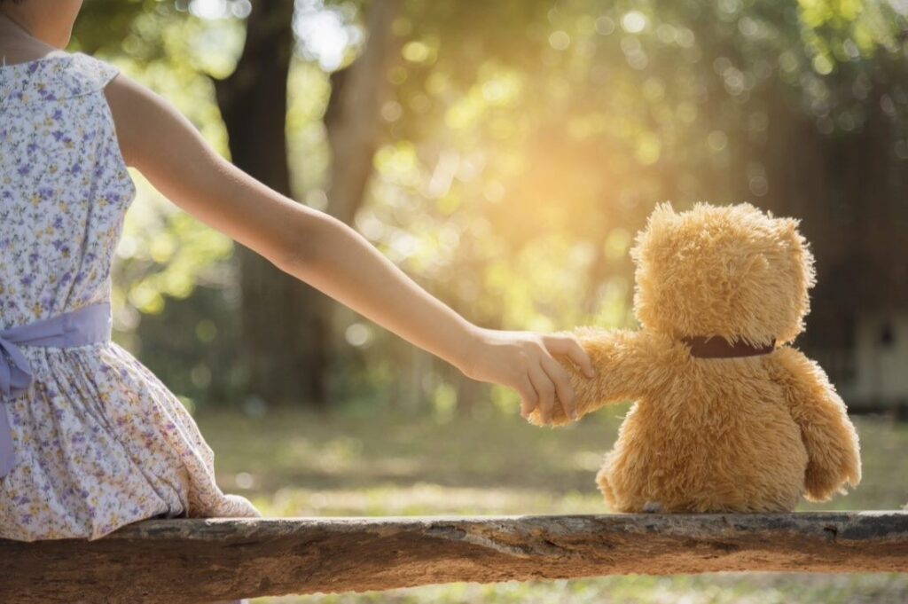  A back shot of a girl holding hands with her stuffed teddy bear as they sit on a log 