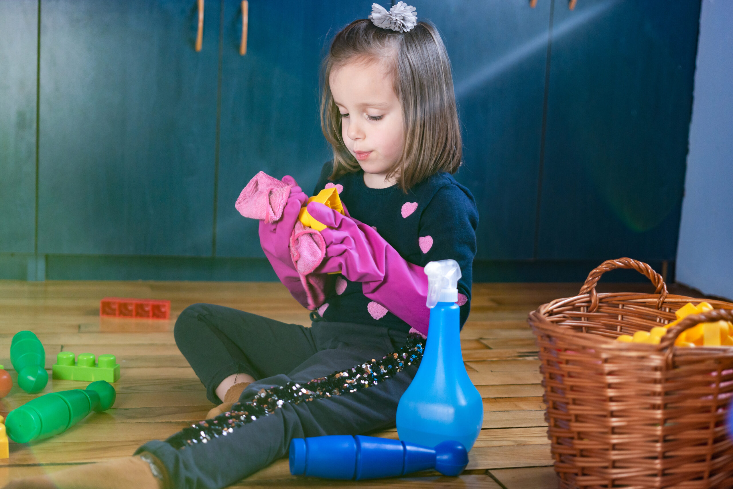 A child sitting on the floor with a spray bottle beside her while she wears oversized gloves to clean her toys