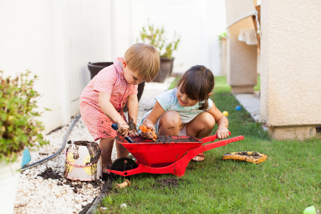 Two toddlers playing with some mud in a small red wheelbarrow