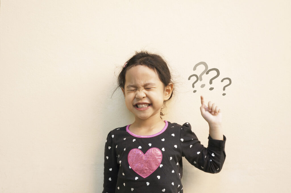A child standing in front of a beige wall while smiling with her eyes closed while pointing to question marks 
