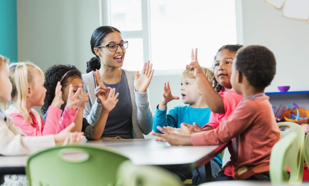  A group of toddlers sitting around a table counting with their hands while their teacher sits in the middle