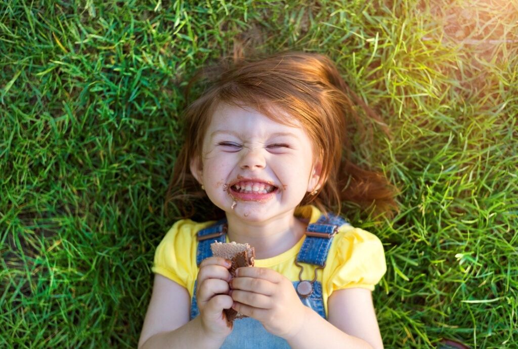 A top shot of a girl lying down on the grass while smiling and eating a snack 
