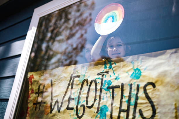 A child in a window holding a rainbow painting while peeking above a poster that says #we got this