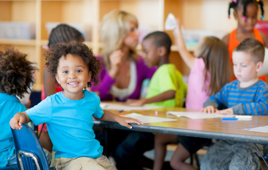 Young child in daycare smiling in classroom with classmates blurred in the background